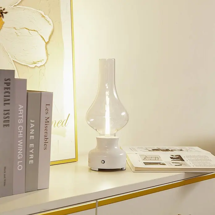 Lindby Tischleuchte Maxentius - L x B x H: 12 x 12 x 30 cm, creme / amber, klar / transparent - Eine Lampe mit weißem Fuß und klarem Glasschirm beleuchtet eine Kommode, umgeben von Büchern und einem Bilderrahmen.