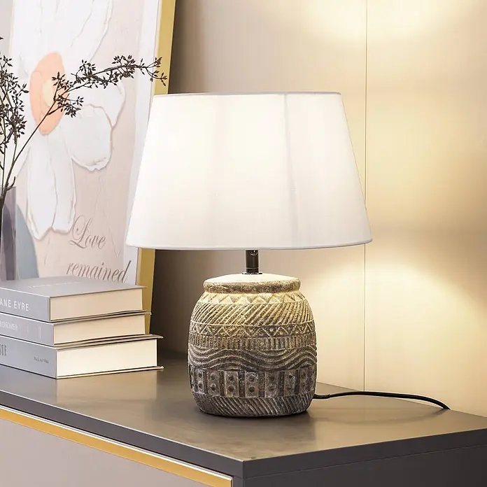 Lindby Tischleuchte Aelith - L x B x H: 30 x 30 x 36 cm, alu / grau / zink, weiß / opal, E27 - Eine dekorative Tischlampe mit geschnitztem, strukturiertem grauem Fuß und weißem Stoffschirm, die einen dunklen Konsolentisch beleuchtet.