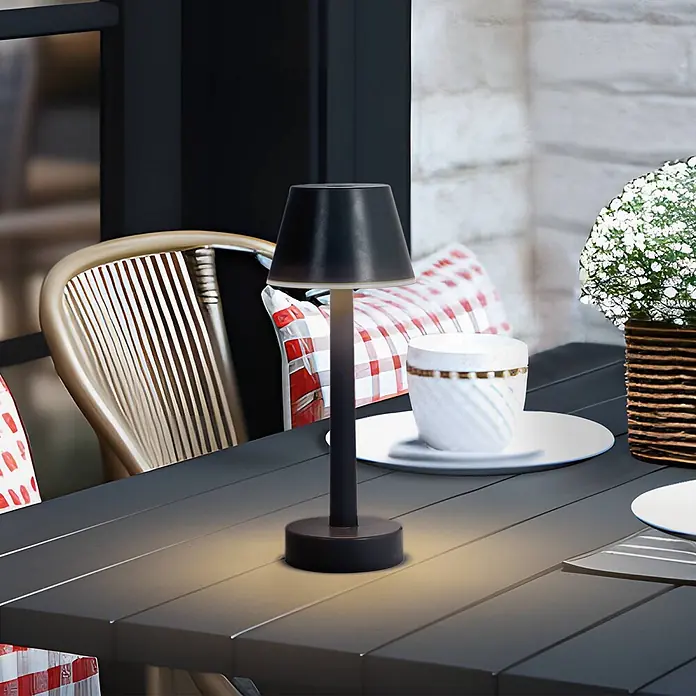 Lindby Tischleuchte Gaja - schwarz, schwarz - Schwarze Tischlampe mit warmem Licht beleuchtet einen Terrassentisch neben Stuhl und Tasse.