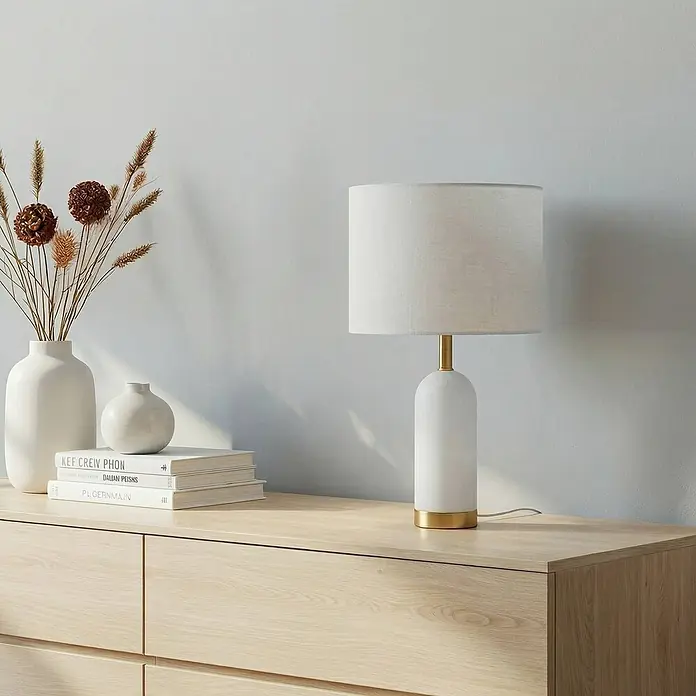 Lindby Tischleuchte Alabaster - alu / grau / zink, weiß / opal, E27 - Weiße und goldene Tischlampe mit hellem Stoffschirm auf einer Holzkommode, neben Vase und Büchern.