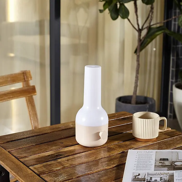 Lindby Tischleuchte Isaline - creme / amber, creme / amber - Weiße und beige tragbare Tischleuchte mit Drehschalter auf einem Holztisch neben einer Tasse und Zeitschrift.