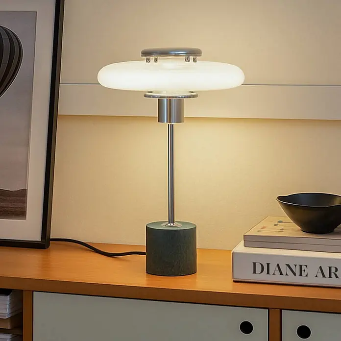 Lindby Tischleuchte Branford - L x B x H: 25 x 25 x 46 cm, grün, weiß / opal, E14 - Eine moderne Tischlampe mit weißem scheibenförmigem Schirm, silbernem Stiel und dunkelgrünem zylindrischem Sockel auf einem Holzschreibtisch.
