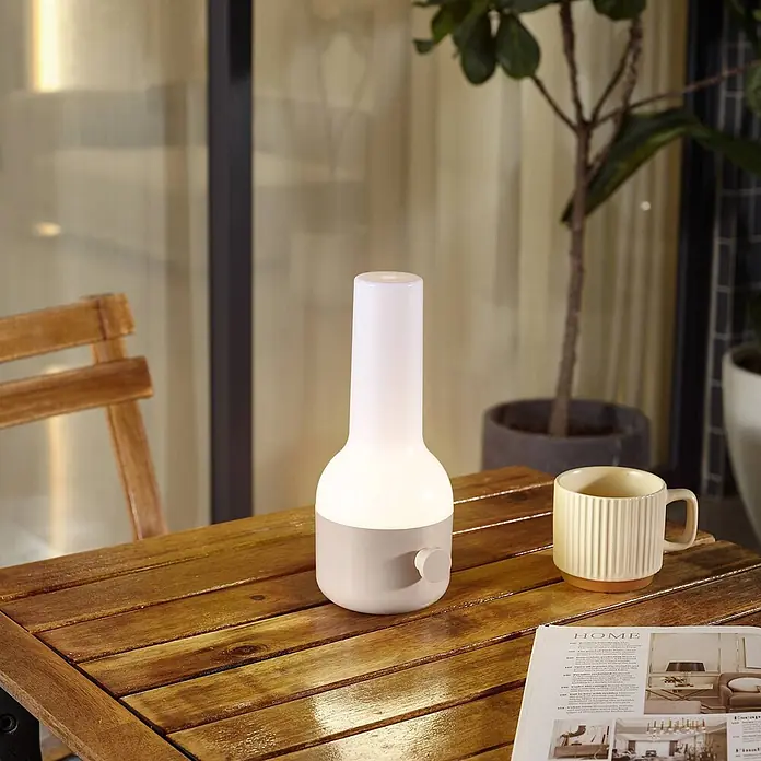 Lindby Tischleuchte Isaline - creme / amber, creme / amber - Schlanke, hellfarbene, tragbare Tischlampe mit sanftem Licht und beigem Sockel auf einem Holztisch im Freien.