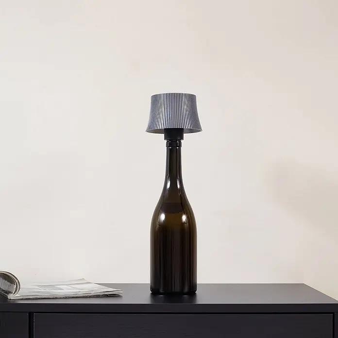 Lindby Tischleuchte Linor - L x B x H: 11,3 x 11,3 x 8,8 cm, alu / grau / zink - Flaschenlampe mit dunkelgrünem Fuß und grauem Plisseeschirm auf einem dunklen Tisch neben einer Zeitschrift.
