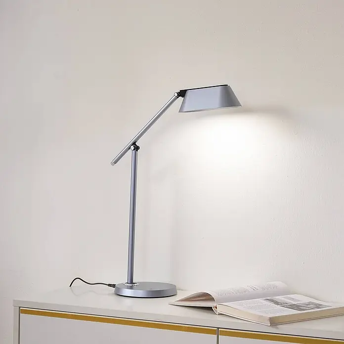 Lindby Tischleuchte Thies - L x B x H: 45,9 x 9,2 x 78,9 cm, alu / grau / zink, alu / grau / zink - Eine silberne, verstellbare Schreibtischlampe beleuchtet ein offenes Buch auf einer weißen Kommode.