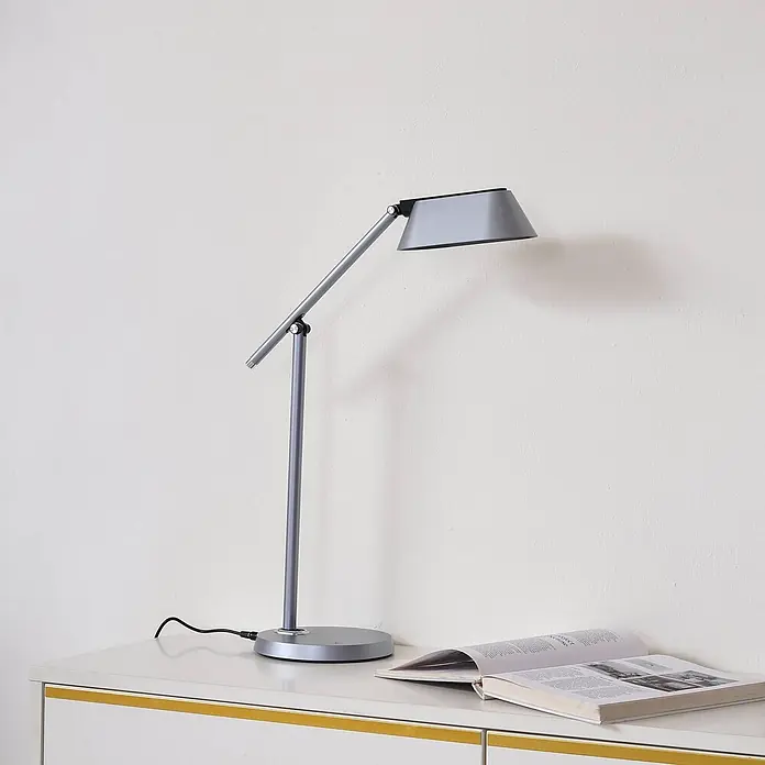 Lindby Tischleuchte Thies - L x B x H: 45,9 x 9,2 x 78,9 cm, alu / grau / zink, alu / grau / zink - Graue Metall-Schreibtischlampe mit verstellbarem Arm und trapezförmigem Schirm, auf einem weißen Tisch neben einem offenen Buch.