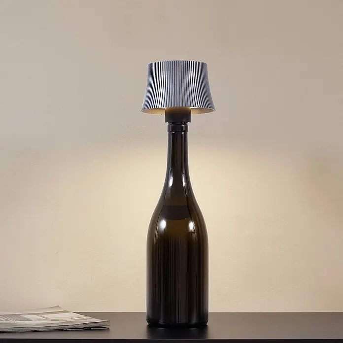 Lindby Tischleuchte Linor - L x B x H: 11,3 x 11,3 x 8,8 cm, alu / grau / zink - Eine beleuchtete Tischlampe mit dunkelgrünem Flaschenfuß und geripptem silbernem Lampenschirm steht auf einer dunklen Oberfläche.