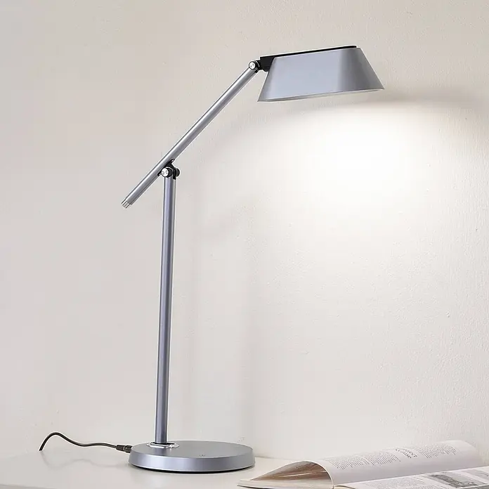Lindby Tischleuchte Thies - L x B x H: 45,9 x 9,2 x 78,9 cm, alu / grau / zink, alu / grau / zink - Eine schlanke, silberne Schreibtischlampe mit verstellbarem Arm und Kopf, die helles Licht auf eine Fläche und ein offenes Buch wirft.