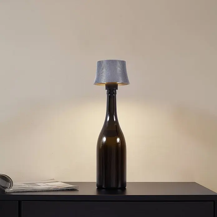Lindby Tischleuchte Linor - L x B x H: 11,3 x 11,3 x 8,8 cm, alu / grau / zink - Flaschenlampe mit dunklem Glasfuß und plissiertem grauem Lampenschirm, die warmes Licht auf einen dunklen Tisch wirft.
