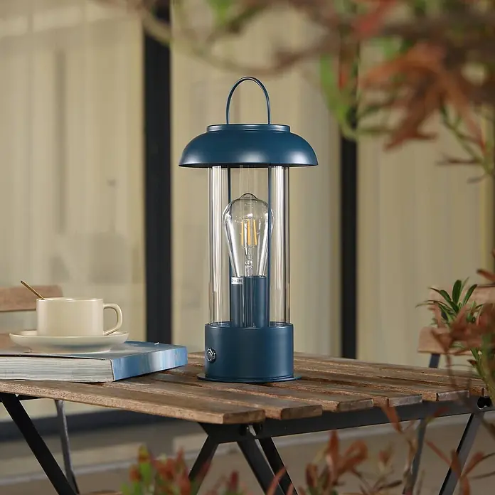Lindby Tischleuchte Yvette - L x B x H: 17 x 17 x 36,4 cm, blau, E27 - Eine dunkel türkisblaue Laternenlampe mit klarem Glasschirm und sichtbarer Glühbirne, auf einem hölzernen Gartentisch.