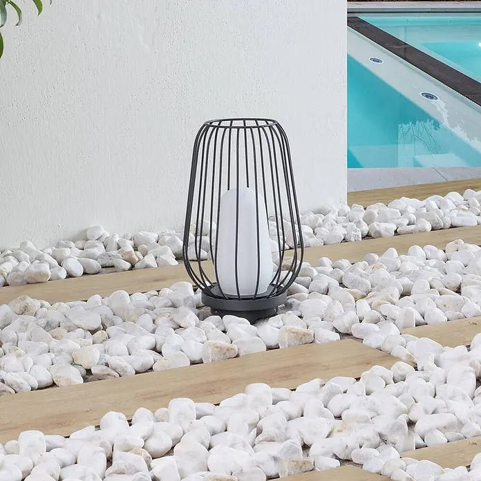 Lucande Tischleuchte Chandan - L x B x H: 20,1 x 20,1 x 32,1 cm, schwarz, schwarz, E27 - Schwarze Metalllaterne mit weißem Lichtmodul, beleuchtet einen Weg aus Holz und weißen Kieselsteinen neben einem Pool.