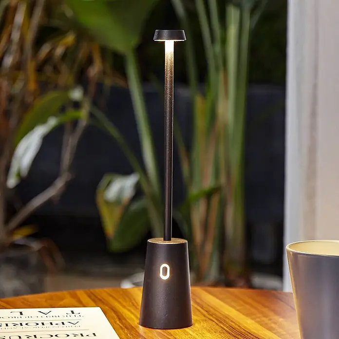 Lindby Tischleuchte Noelani - L x B x H: 5,5 x 5,5 x 30 cm, schwarz, schwarz - Eine schlanke, dunkle Metall-Tischlampe mit beleuchteter Oberseite und Ziffer '0' auf dem Sockel steht auf einem Holztisch neben einem Buch und einer Tasse.