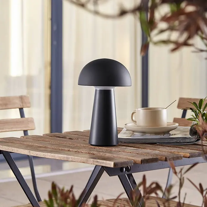 Lindby Tischleuchte Zyre - schwarz, schwarz - Schwarze pilzförmige Tischlampe auf einem rustikalen Holztisch im Freien.