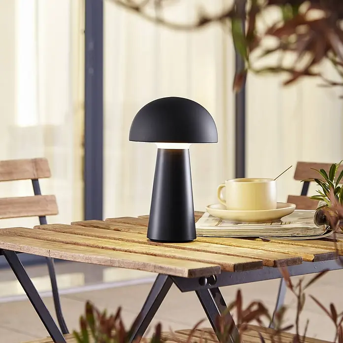 Lindby Tischleuchte Zyre - schwarz, schwarz - Eine matte schwarze Pilz-Tischlampe mit warmem Licht steht auf einem Holz-Außentisch neben einer gelben Tasse.