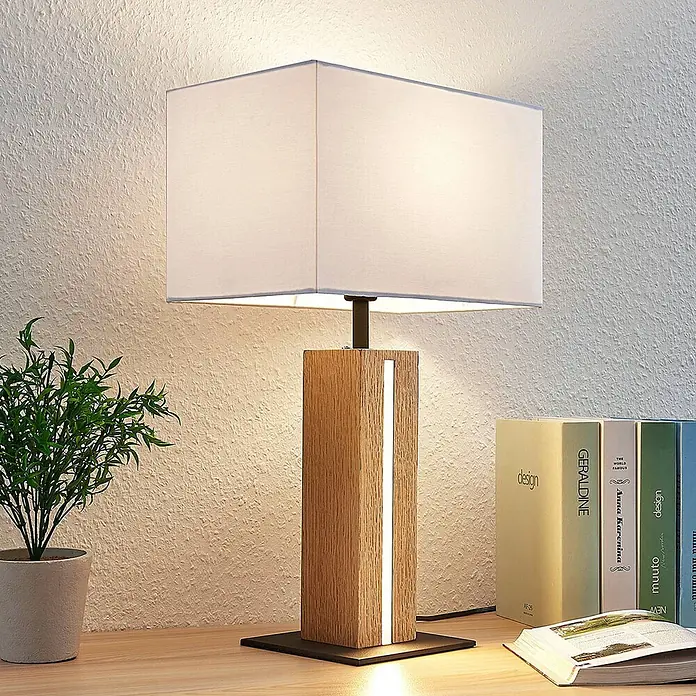 Lindby Tischleuchte Garry - L x B x H: 32 x 20 x 52,5 cm, Holz hell, weiß / opal, E27 - Eine Tischlampe mit rechteckigem weißem Stoffschirm und einem Holzsockel mit integrierter vertikaler LED-Leiste auf einer Holzoberfläche.