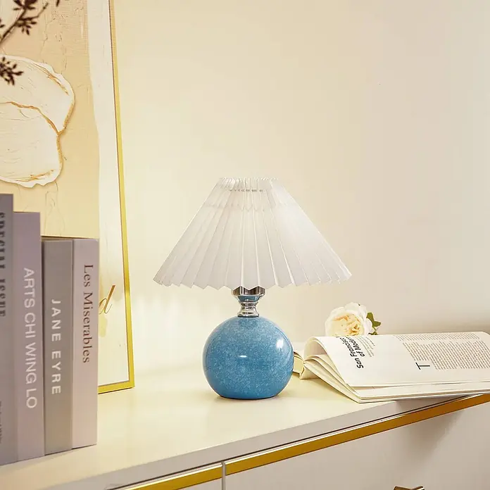 Lindby Tischleuchte Kerimi - L x B x H: 24 x 24 x 29 cm, blau, blau, E27 - Eine blaue kugelförmige Tischlampe mit weißem plissiertem Schirm, auf einer Oberfläche neben Büchern und einer Blume.