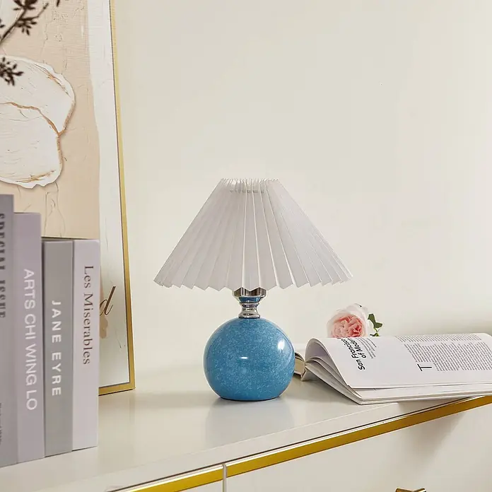 Lindby Tischleuchte Kerimi - L x B x H: 24 x 24 x 29 cm, blau, blau, E27 - Blaue kugelförmige Keramik-Tischlampe mit weißem Plisseeschirm auf einer weißen Oberfläche mit Büchern und Magazin.