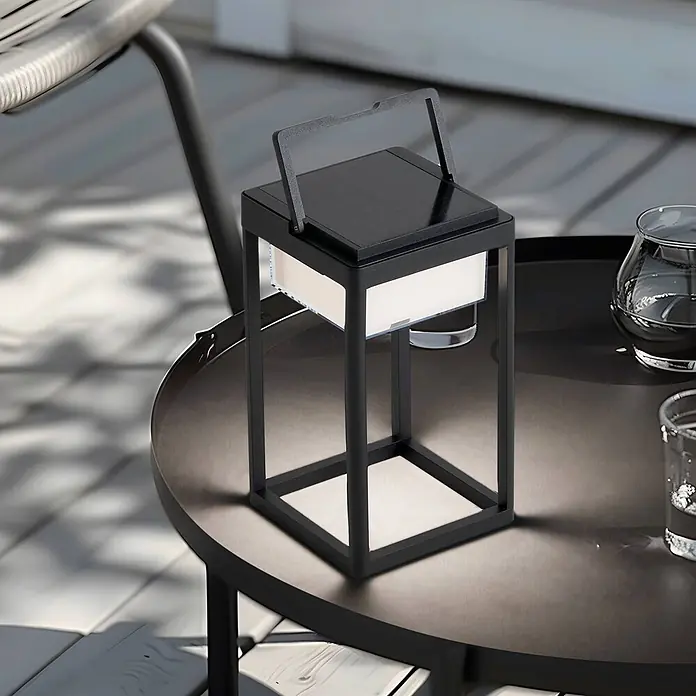 Lucande Tischleuchte Tilena - L x B x H: 18,5 x 18,5 x 32 cm, schwarz - Schwarze, rechteckige Solarlaterne mit Griff, leuchtet von den Seiten, steht auf einem Gartentisch.