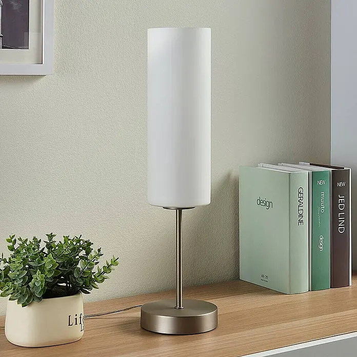 Lindby Tischleuchte Felice - L x B x H: 10 x 10 x 46 cm, alu / grau / zink, weiß / opal, E27 - Eine Tischlampe mit zylindrischem weißem Schirm und gebürstetem Metallsockel steht auf einem Holzschreibtisch neben einer Pflanze und Büchern.