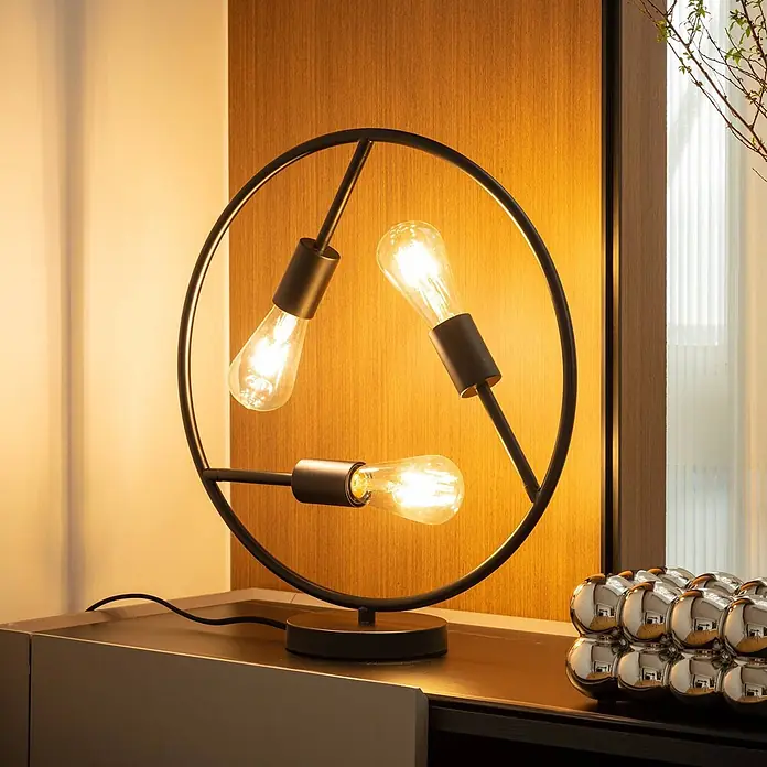 Lucande Tischleuchte Linnard - L x B x H: 45 x 45 x 50 cm, schwarz, E27 - Moderne schwarze kreisförmige Tischlampe mit drei sichtbaren Edison-Glühbirnen auf einer dunklen Kommode, die eine warme Holzwand beleuchtet.