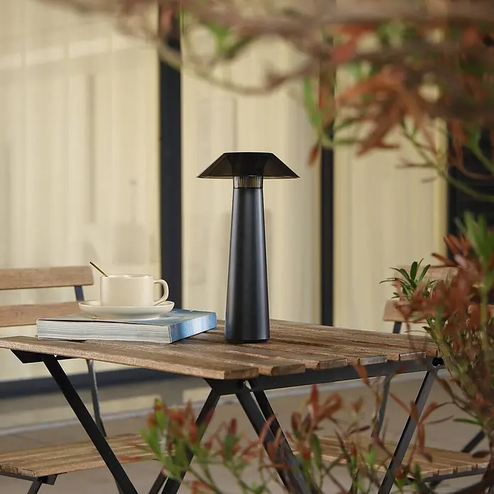 Lindby Tischleuchte Gomba - schwarz - Elegante schwarze tragbare Lampe mit dunklem Schirm auf einem Holztisch neben einer Tasse und einem Buch.
