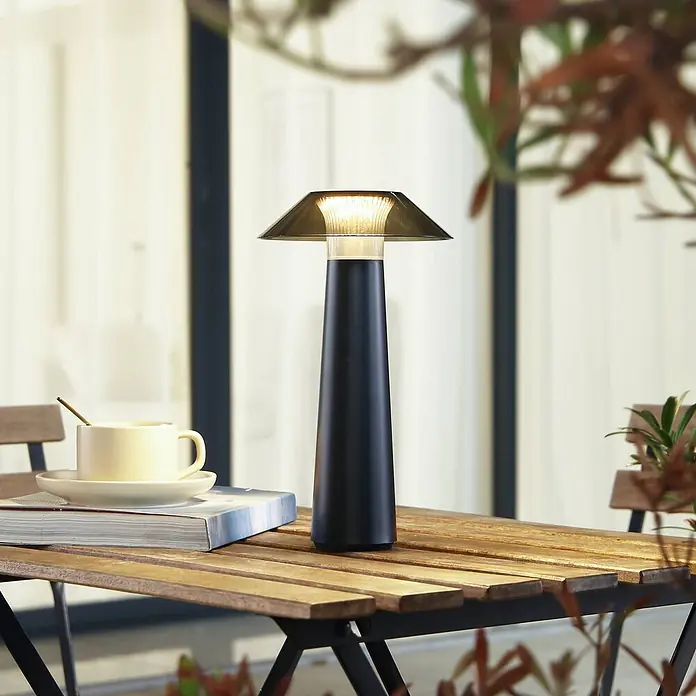 Lindby Tischleuchte Gomba - schwarz - Eine schwarze konische Tischlampe mit rauchig-transparenter Haube, warm leuchtend, auf einem hölzernen Gartentisch neben einer Kaffeetasse und einem Buch.