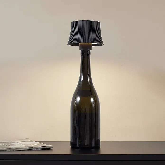 Lindby Tischleuchte Linor - schwarz - Eine Tischlampe aus einer dunkelgrünen Flasche mit schwarzem plissiertem Lampenschirm, die warmes Licht auf einen dunklen Tisch wirft.