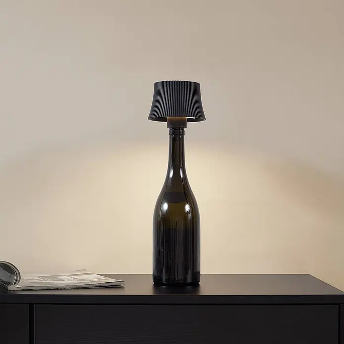 Lindby Tischleuchte Linor - schwarz - Dunkle Glasflaschenlampe mit schwarzem Plisseeschirm auf dunklem Holztisch, beleuchtet helle Wand.