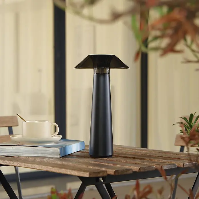 Lindby Tischleuchte Gomba - schwarz - Eine schwarze konische Tischlampe mit dunklem, transparentem Trapezschirm steht auf einem Holzlamellentisch im Freien, neben einer Kaffeetasse und einem Buch.
