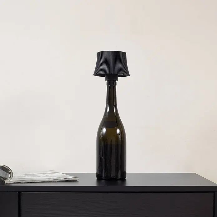 Lindby Tischleuchte Linor - schwarz - Dunkelgrüne Flaschenlampe mit schwarzem Plisseeschirm auf dunklem Holztisch.