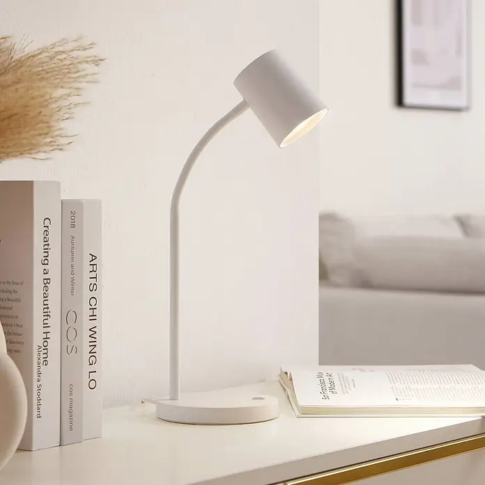 Lindby Tischleuchte Radka - L x B x H: 14,2 x 14,2 x 40,5 cm, weiß / opal, weiß / opal, GU10 - Eine weiße Tischlampe mit flexiblem Arm, die warmes Licht auf ein offenes Buch und weitere Bücher auf einem hellen Schreibtisch wirft.