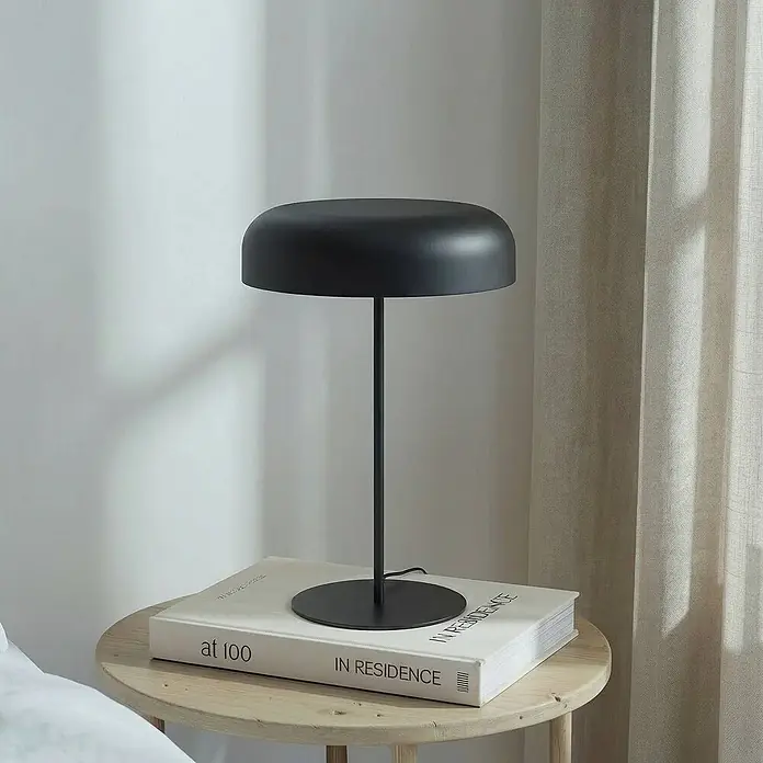 Lindby Tischleuchte Edion - schwarz, schwarz, E14 - Eine schwarze, moderne Tischlampe steht auf einem hellen Holznebentisch, unter ihrer Basis liegt ein großes weißes Buch.