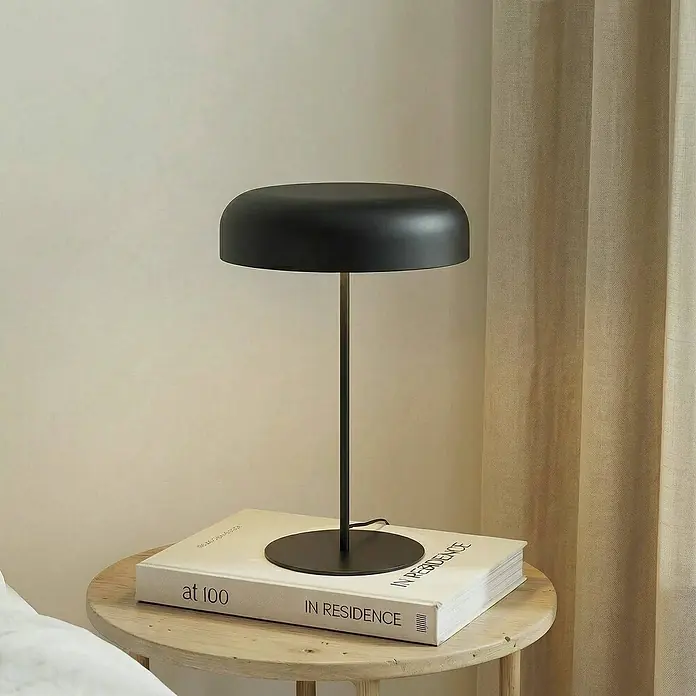 Lindby Tischleuchte Edion - schwarz, schwarz, E14 - Eine schwarze, minimalistische Tischleuchte mit gewölbtem Schirm und dünnem Stiel, auf einem Buch auf einem hellen Holztischchen.