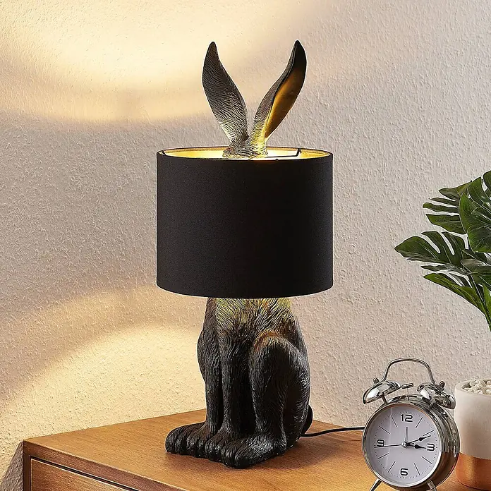 Lindby Tischleuchte Lorentina - L x B x H: 24 x 24 x 56 cm, schwarz, E27 - Hasenförmige Tischlampe mit dunkler Oberfläche und schwarzem Schirm, die einen Nachttisch beleuchtet.