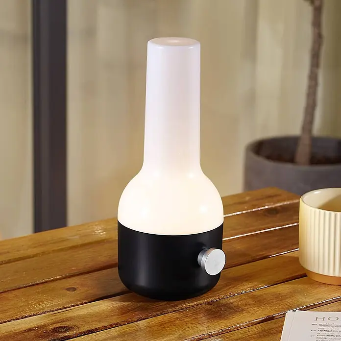 Lindby Tischleuchte Isaline - schwarz, weiß / opal - Eine moderne schwarz-weiße Tischlampe mit silbernem Drehknopf auf einem Holztisch, die Umgebungslicht spendet.