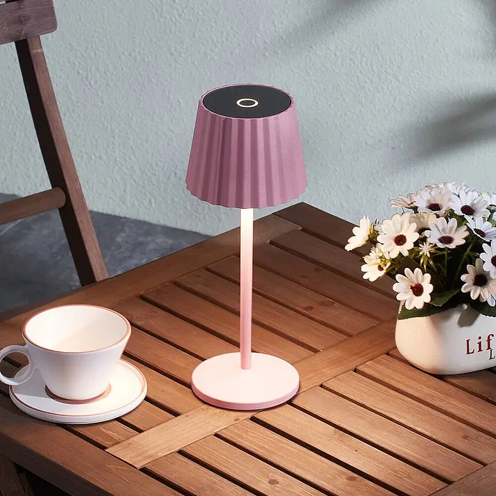 Lindby Tischleuchte Esali - L x B x H: 11 x 11 x 28,5 cm, violett / pink / rose, violett / pink / rose - Rosa Tischlampe mit plissiertem Schirm, die einen Holztisch im Freien beleuchtet.