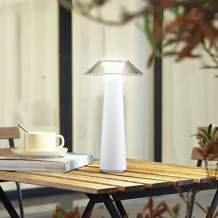 Lindby Tischleuchte Gomba - weiß / opal - Weiße, konische Lampe mit transparenter, facettierter Oberseite auf einem Holztisch im Freien mit einer Tasse.