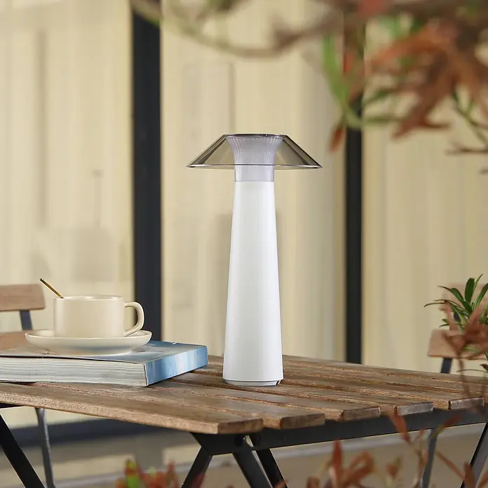 Lindby Tischleuchte Gomba - weiß / opal - Weiße tragbare Tischlampe mit transparentem Schirm auf einem Holztisch im Freien, neben Tasse und Buch.