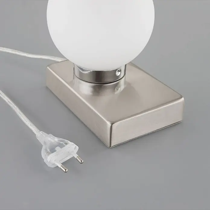 Lindby Tischleuchte Naomi - alu / grau / zink, weiß / opal, E14 - Nahaufnahme einer Tischlampe mit kugelförmigem weißen Glasschirm, gebürstetem Metallsockel und transparentem Netzkabel mit Eurostecker.