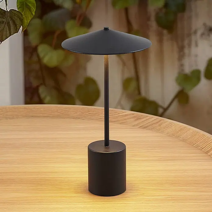 Lindby Tischleuchte Ayva - schwarz, schwarz - Schwarze minimalistische Tischlampe mit kegelförmigem Schirm, die eine Holzoberfläche vor unscharfem Grün beleuchtet.