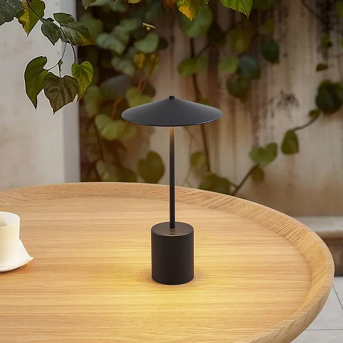 Lindby Tischleuchte Ayva - schwarz, schwarz - Schwarze Metall-Tischlampe mit scheibenförmigem Schirm, wirft warmes Licht auf einen hellen Holztisch.