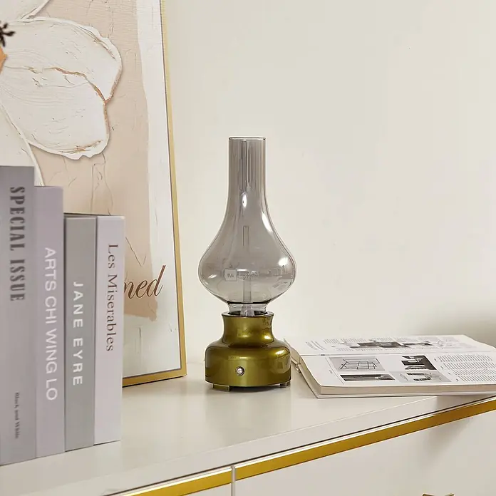 Lindby Tischleuchte Maxentius - L x B x H: 12 x 12 x 30 cm, grün, alu / grau / zink - Eine Tischlampe im Antik-Stil mit goldfarbenem Metallsockel und rauchgrauem Glasschirm, auf einem weißen Sideboard neben Büchern.