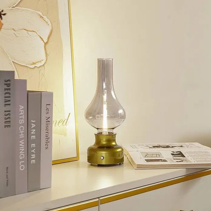 Lindby Tischleuchte Maxentius - L x B x H: 12 x 12 x 30 cm, grün, alu / grau / zink - Vintage olivgoldene Tischlampe mit getöntem Glasschirm auf einer weißen Kommode neben Büchern, warmes Licht ausstrahlend.