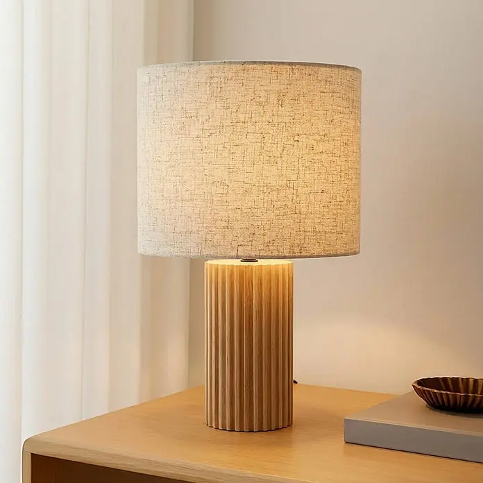 Lindby Tischleuchte Kimiko - L x B x H: 25 x 25 x 38 cm, Holz hell, E27 - Tischlampe mit gerilltem zylindrischem Holzfuß und beigem Leinenschirm, die einen Holznebentisch beleuchtet.