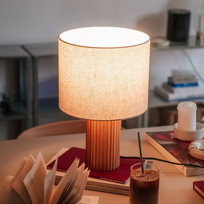 Lindby Tischleuchte Kimiko - L x B x H: 25 x 25 x 38 cm, Holz hell, E27 - Eine Tischlampe mit hellem Leinenschirm und geriffelter Holzbasis beleuchtet einen Schreibtisch mit Büchern und einem Getränk.