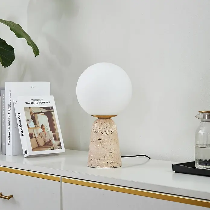 Lindby Tischleuchte Kirstan - creme / amber, weiß / opal, E27 - Tischlampe mit weißem Glaskugel-Schirm und konischem, hellem Steinsockel auf einer Kommode.