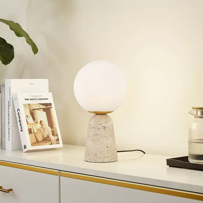 Lindby Tischleuchte Kirstan - creme / amber, weiß / opal, E27 - Eine Tischlampe mit kugelförmigem weißem Glasschirm und strukturiertem, hellem Travertinsockel auf einer weißen Kommode.
