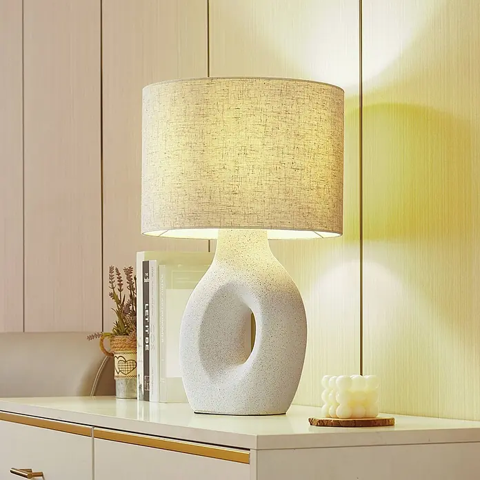 Lindby Tischleuchte Thalassia - weiß / opal, weiß / opal, E27 - Moderne Tischlampe mit beigem Stoffschirm und weißem Keramikfuß mit Ausschnitt, spendet warmes Licht auf einem Konsolentisch.