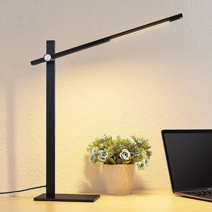 Lucande Tischleuchte Wasko - L x B x H: 45,5 x 12 x 47 cm, schwarz, schwarz - Moderne schwarze LED-Schreibtischlampe beleuchtet einen Holztisch neben einer Pflanze und einem Laptop.
