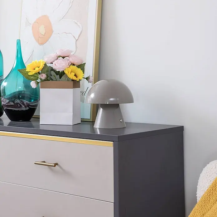 Lindby Tischleuchte Nevijo - alu / grau / zink, alu / grau / zink - Graue, pilzförmige Tischlampe auf einer dunkelgrauen Kommode mit Schubladen, neben Dekorationselementen.